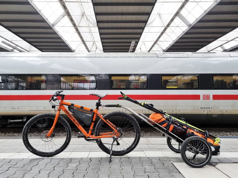 Fahrrad mit Ski und Gepäck auf einem Anhänger am Bahnsteig.