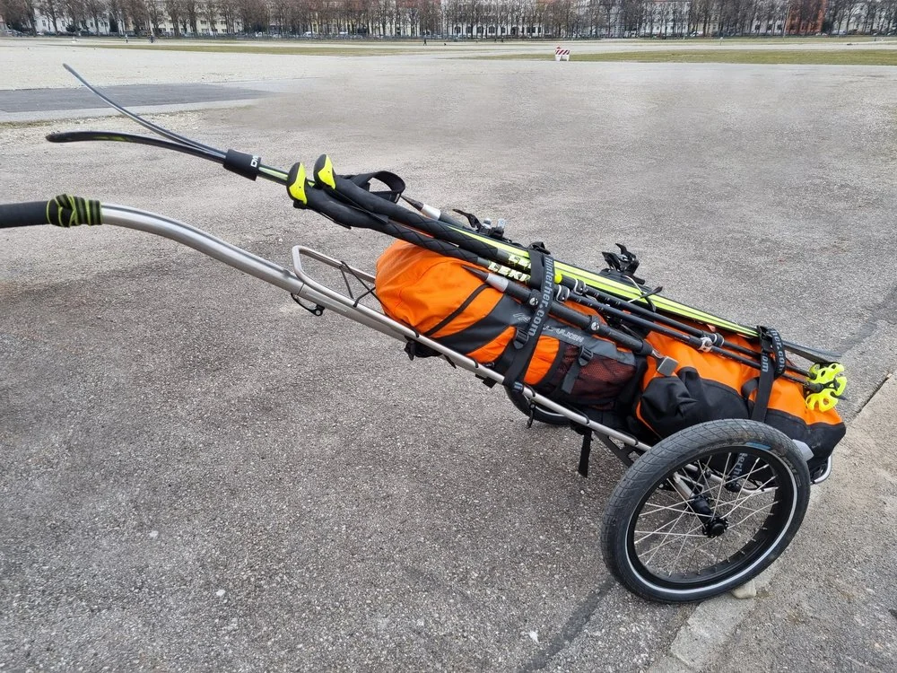 Ski, Stöcke und Gepäck sind mit Gurten auf dem zweirädrigen Hinterher-Trolley befestigt.